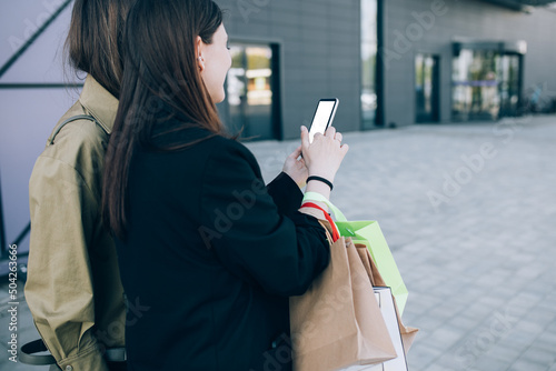 Two happy women friends holding bags enjoying shopping, using smartphone. photo