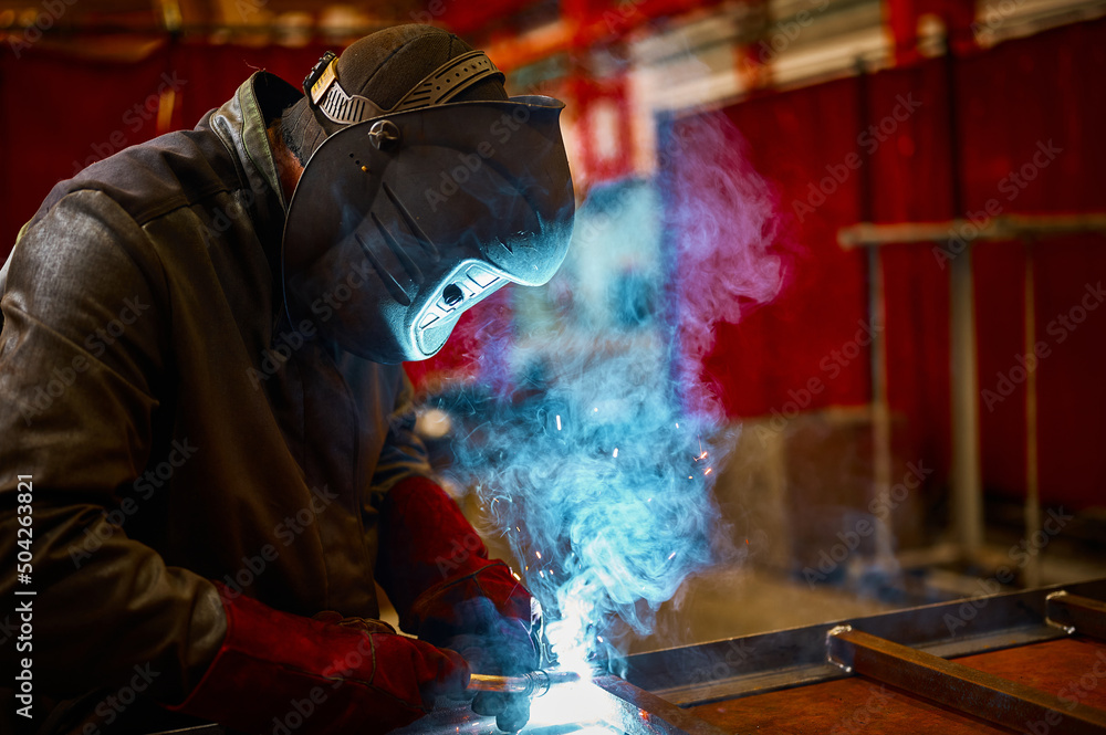Skilled worker in protective mask welds metal part at plant Skilled worker in protective mask welds metal part at plant