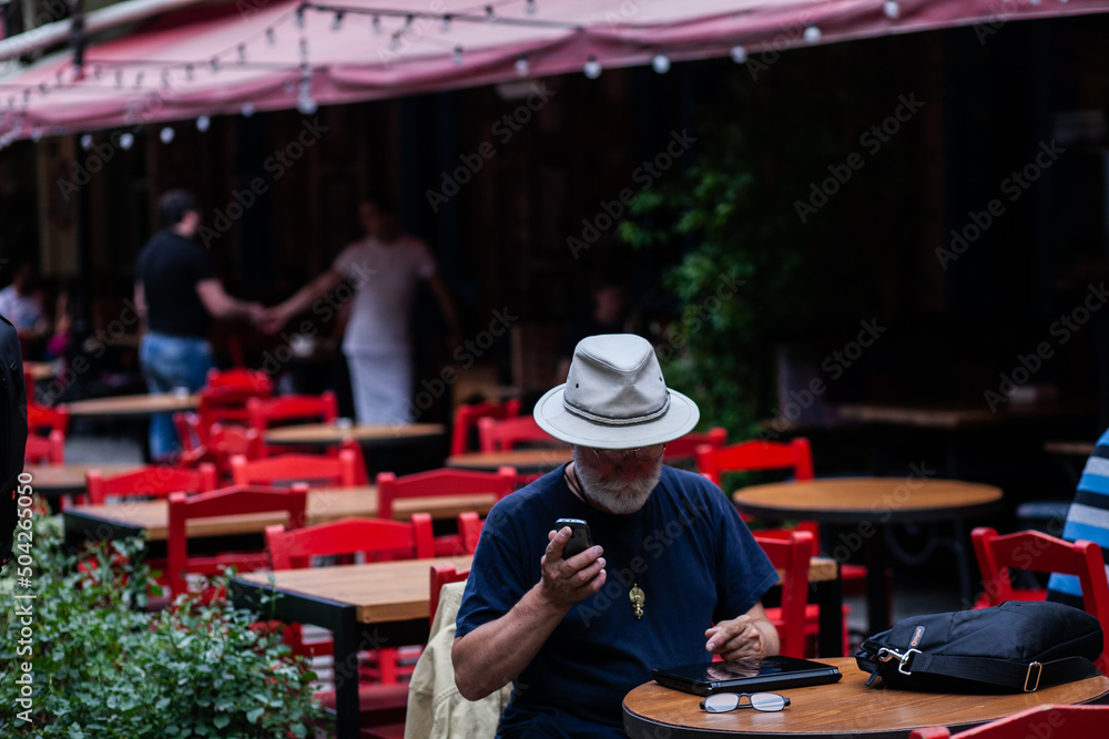 a man in a white hat is sitting in a street cafe against the background of red chairs, holding a phone, glasses and a bag on the table, medium plan, front and side views