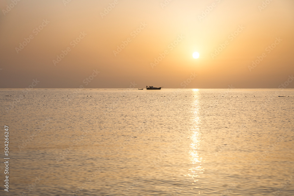 Small ship with boat and orrange disk of the sun setting over the sea during sunset on the Mediterranean sea, Turkey, Antalya Province