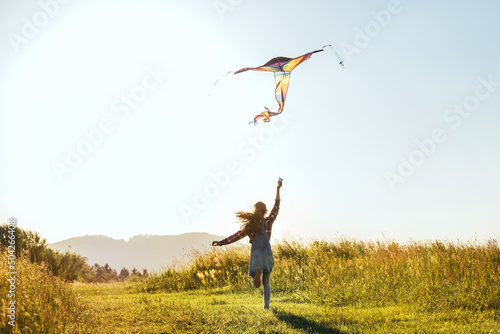 Long-haired Girl with flying a colorful kite on the high grass meadow in the mountain fields. Happy childhood moments or outdoor time spending concept image. #504266408
