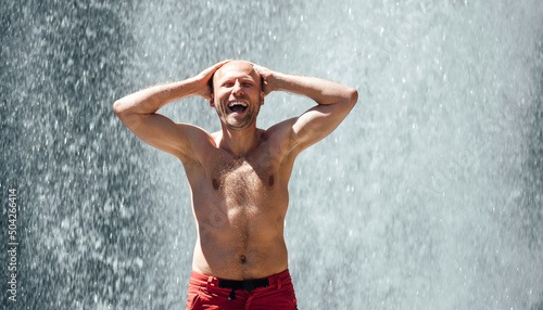 Middle-aged shirtless sincerely laughing man standing under the mountain river waterfall and enjoying the splashing Nature power. Fit people, trekking, and a natural beauty concept image. © Soloviova Liudmyla