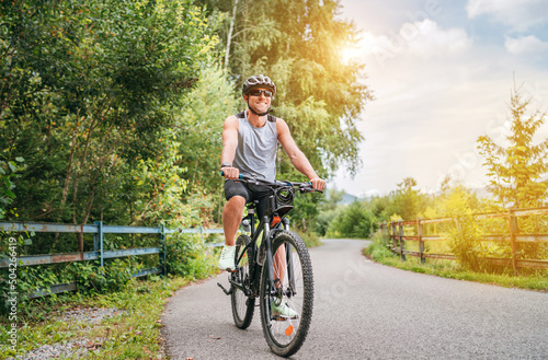 Portrait of a happy smiling man dressed in cycling clothes, helmet and sunglasses riding a bicycle on the asphalt out-of-town bicycle path. Active sporty people concept image.