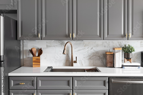 A kitchen sink detail shot with grey cabinets, a white marble countertop and backsplash, and decorations. photo