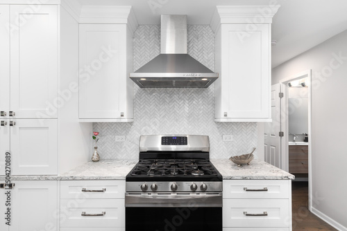 A stainless steel oven and hood in a white kitchen with marble countertops, a herringbone backsplash, and a bathroom down the hall. 