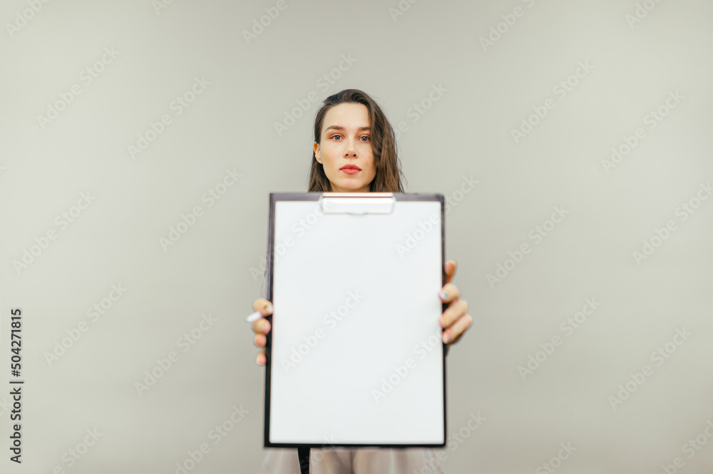 Beautiful female office worker holds a paper tablet with a white sheet and shows to the camera isolated on a beige background. Beautiful female office worker holds a paper tablet with a white sheet and shows to the camera isolated on a beige background.