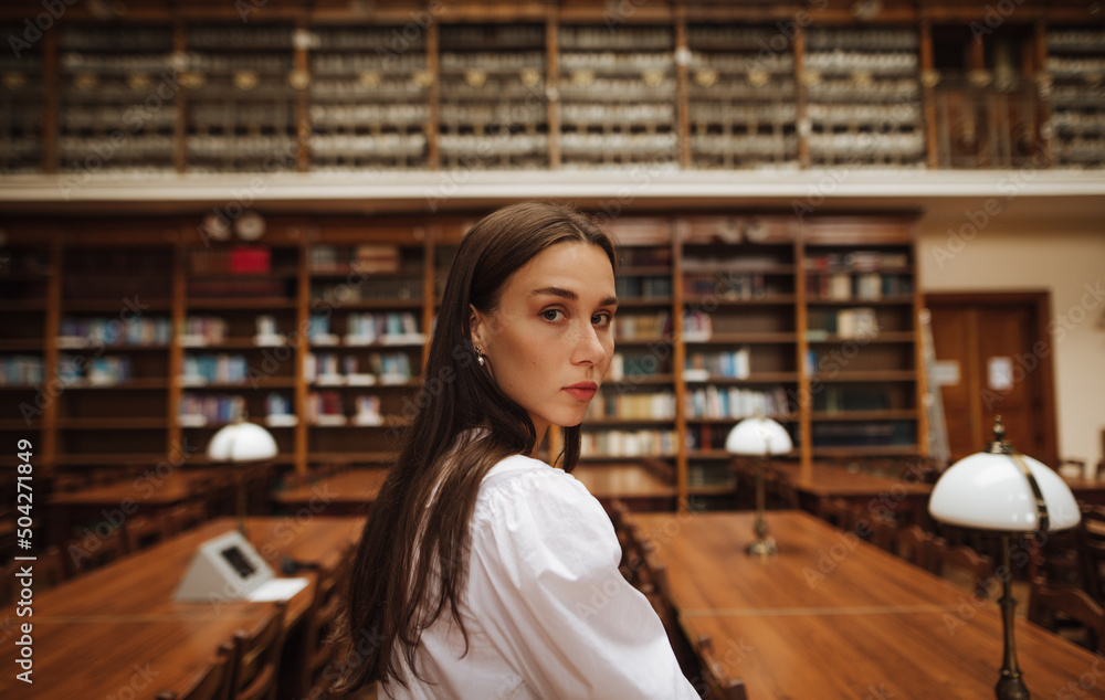 Portrait of a beautiful girl in a public library posing at camera on a background of shelves with books. Portrait of a beautiful girl in a public library posing at camera on a background of shelves with books.