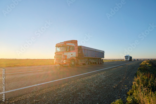 Semi-truck with tipping cargo trailer transporting sand from quarry driving on highway hauling goods in evening. Delivery transportation and logistics concept