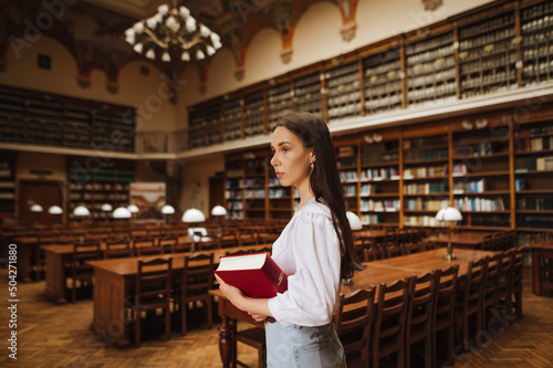 Portrait of a beautiful student in a white blouse studying in the university library, standing with a book in hand and looking away.