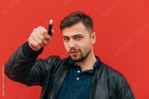 Funny adult man with a beard smokes an electronic cigarette against a red wall and shows an iqos to the camera.