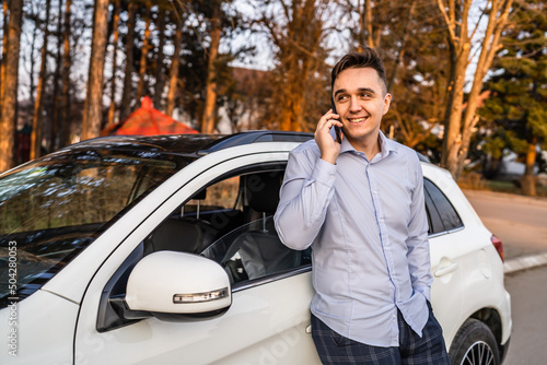 One man young adult businessman using mobile phone while standing in front of his car outdoor making a phone-call talking happy smile in day wearing shirt looking busy real people copy space