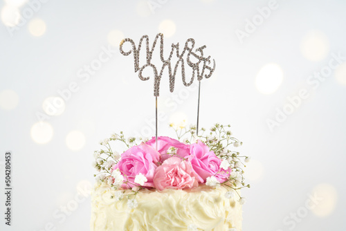 Wedding 2 Tiered Cake with Mr and Mrs topper. Styled with fresh pink roses and gypsophila flowers, against a white background with bokeh party fairy lights. Close up.