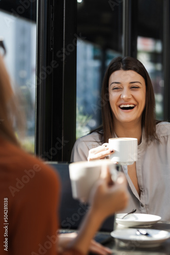 Two young business women in a cafe having one on one meeting. Friends after work talking gossiping and having coffee at a window table on a sunny day..