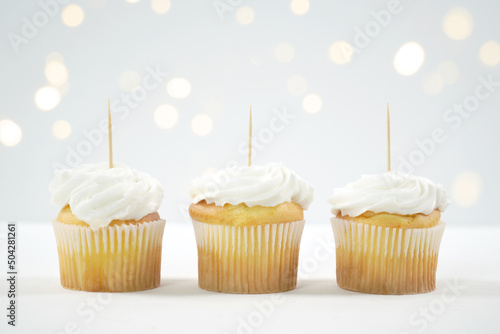Three Vanilla Cupcakes Topper Mockup. Styled against a white background with bokeh party fairy lights. Copy space for your design here.