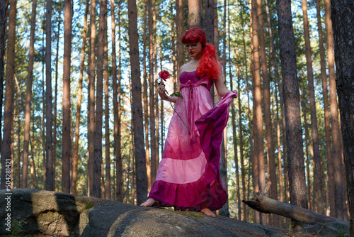 a girl with red hair and a dress with a rose in her hands is resting in a fabulous forest.