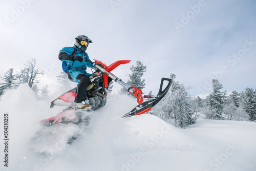 Motorcyclist jumping on snowbike in snow splashes in deep snow dust on winter day