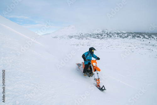 Snowbike rider going down steep slope in mountain valleyr. Modify dirt bike with snow splashes and trail