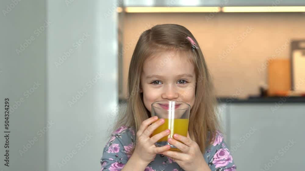 Portrait of little girl looks at camera, laughs and fun drinks orange juice. Happy child drinks juice on kitchen background Portrait of little girl looks at camera, laughs and fun drinks orange juice. Happy child drinks juice on kitchen background