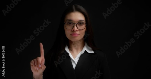No sign gesture. Portrait of serious young businesswoman raising forefinger up saying no. A brunette with long hair wearing glasses and business clothes does not agree. Isolated on a black background