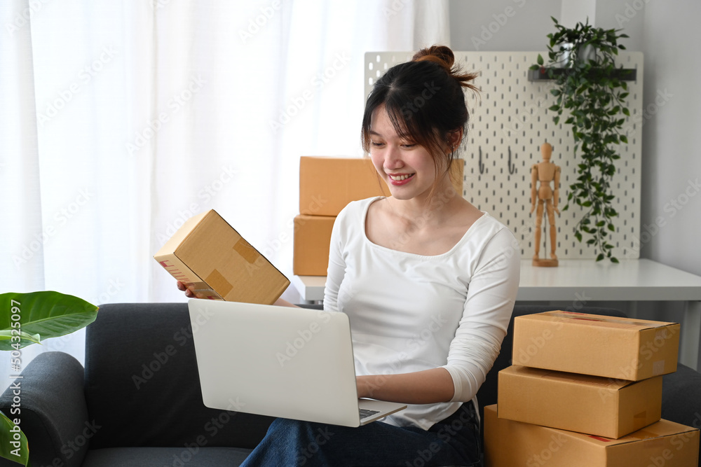 Smiling female freelancer checking product purchase order on laptop and preparing parcel for delivery. Smiling female freelancer checking product purchase order on laptop and preparing parcel for delivery.