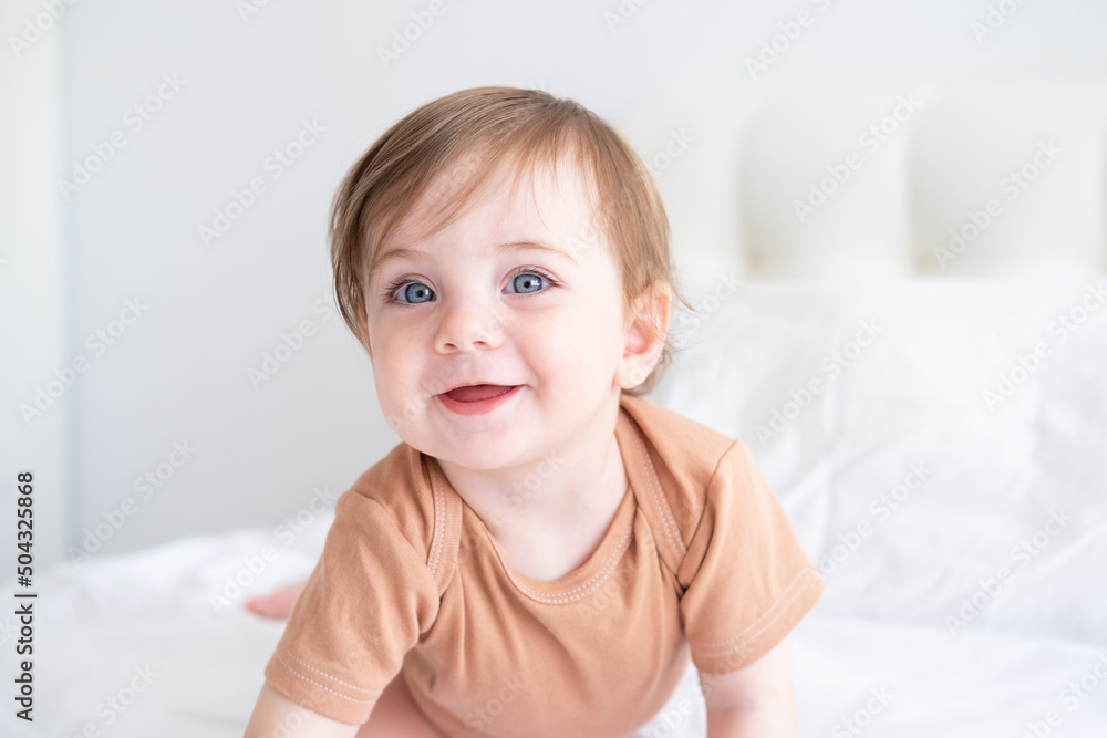 portrait of cute baby girl 10 months with blue eyes in brown bodysuit on white bedding on bed