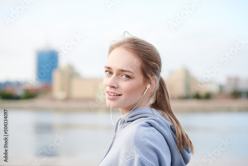 Portrait of smiling attractive girl in hoodie listening to music in earphone against city riverside background