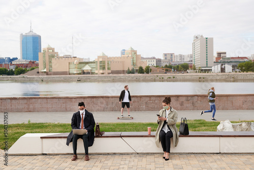 Modern business people in facial masks sitting on bench on embankment and working with devices outside