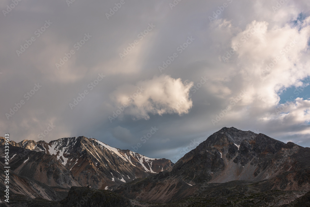 Dramatic mountain landscape with high mountain range with sharp rocky pinnacle under clouds of sunset color in gloomy sky. Dark atmospheric scenery with large mountains with snow at changeable weather