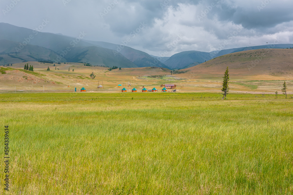 Scenic view from sunlit grassy steppe to small houses on background of green high mountain range in rainy clouds. Dramatic landscape with meadow and small lodges in mountains at changeable weather.