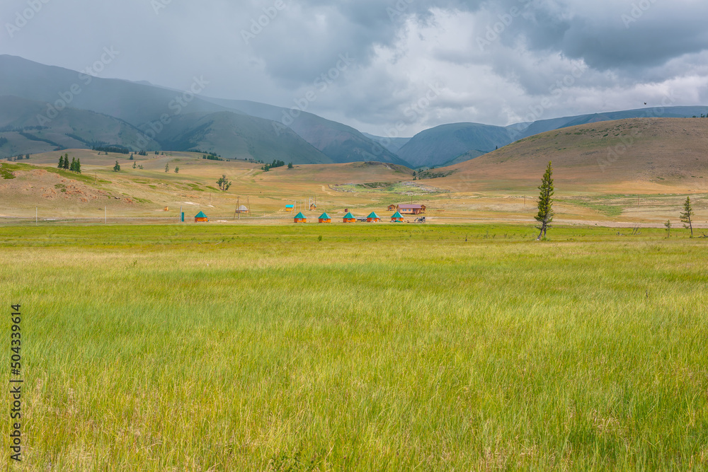 Scenic view from sunlit grassy steppe to small houses on background of green high mountain range in rainy clouds. Dramatic landscape with meadow and small lodges in mountains at changeable weather. Scenic view from sunlit grassy steppe to small houses on background of green high mountain range in rainy clouds. Dramatic landscape with meadow and small lodges in mountains at changeable weather.