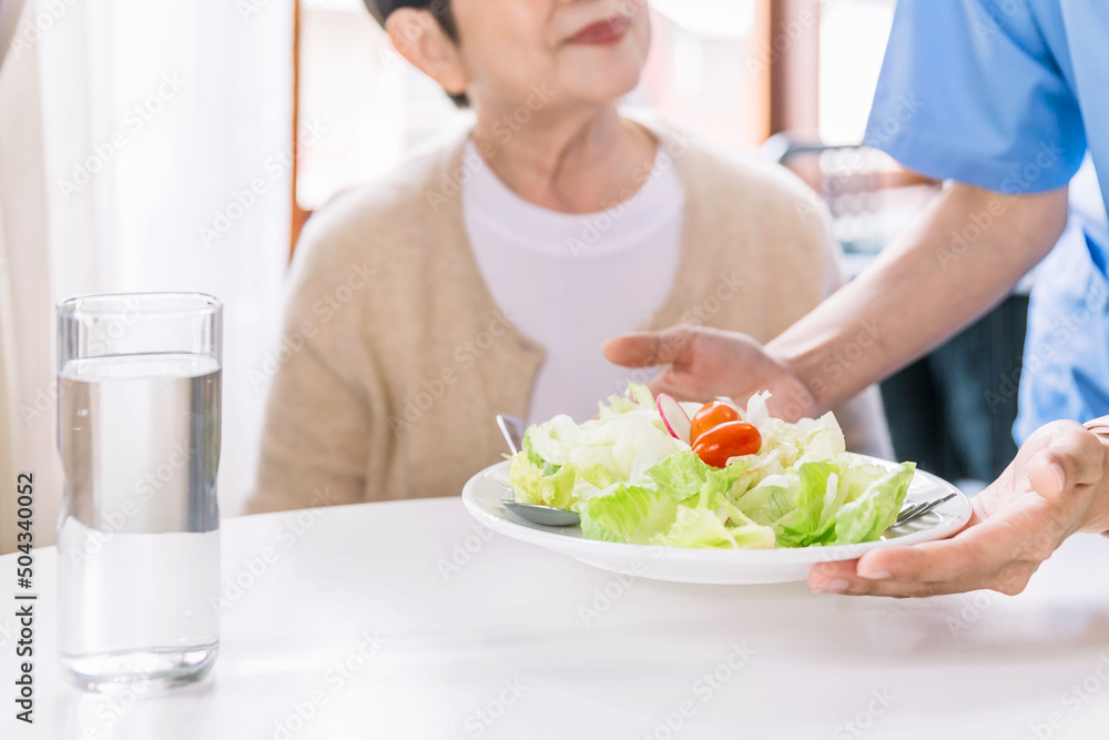 Close up hands of caregiver wearing medical scrubs caring to senior Asian woman by serving a vegetarian salad eating meals at home. Caregiver visit at home. Home health care and nursing home concept. Close up hands of caregiver wearing medical scrubs caring to senior Asian woman by serving a vegetarian salad eating meals at home. Caregiver visit at home. Home health care and nursing home concept.