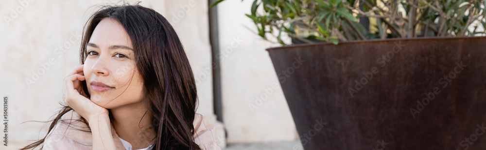 brunette woman holding hand near face while looking away near huge flowerpot outdoors, banner. brunette woman holding hand near face while looking away near huge flowerpot outdoors, banner.