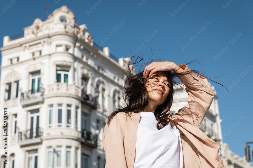 low angle view of brunette woman fixing hair while standing on wind in city.