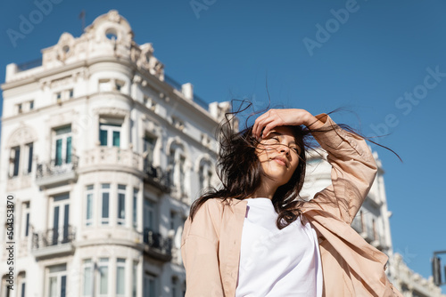 low angle view of brunette woman fixing hair while standing on wind in city.