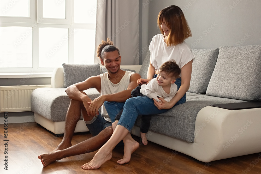 Happy, modern, multiracial family with a little boy in cozy home clothes sit on a gray sofa near a large window, laugh Happy, modern, multiracial family with a little boy in cozy home clothes sit on a gray sofa near a large window, laugh