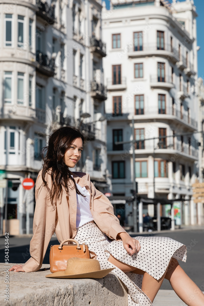stylish brunette woman sitting on street near straw hat and handbag.