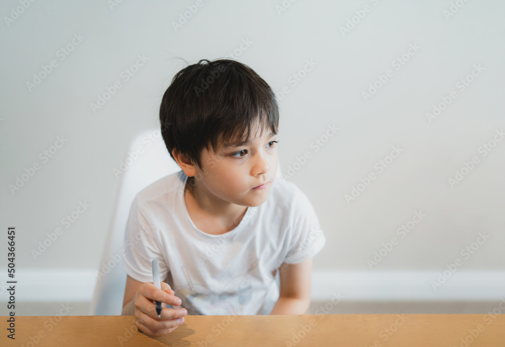 Portrait Young boy holding black pen looking deep in thought, School Kid looking out with thinking face. Child with curious face sitting alone on table Portrait Young boy holding black pen looking deep in thought, School Kid looking out with thinking face. Child with curious face sitting alone on table