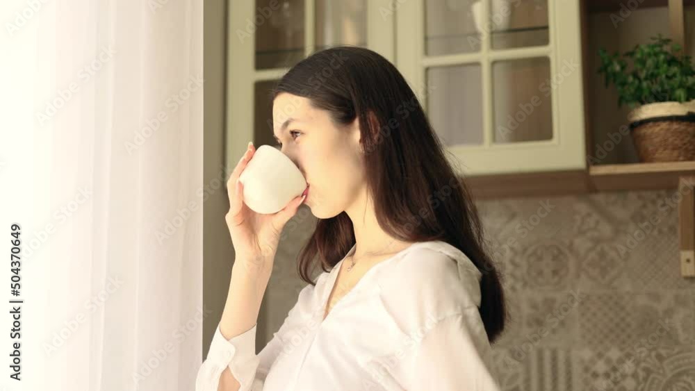 Young beautiful woman looks out the window, drinks a hot drink of tea or coffee, the rays of the rising sun fall on her face. Beautiful morning. Young beautiful woman looks out the window, drinks a hot drink of tea or coffee, the rays of the rising sun fall on her face. Beautiful morning.