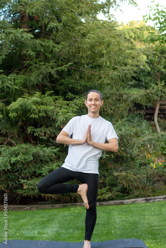 Young man smiling trying and learning how to do a yoga pose in the garden for fun wearing sportswear
