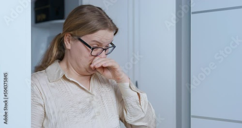 Allergic curvy woman sneezes after cleaning dust in room. Blonde housewife in black-rimmed glasses stands holding on wardrobe at home close view photo