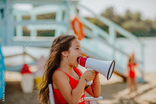 Pretty teen girl in red bikini posing with megaphone sitting on white summer chair at the beach against blue lifeguard tower