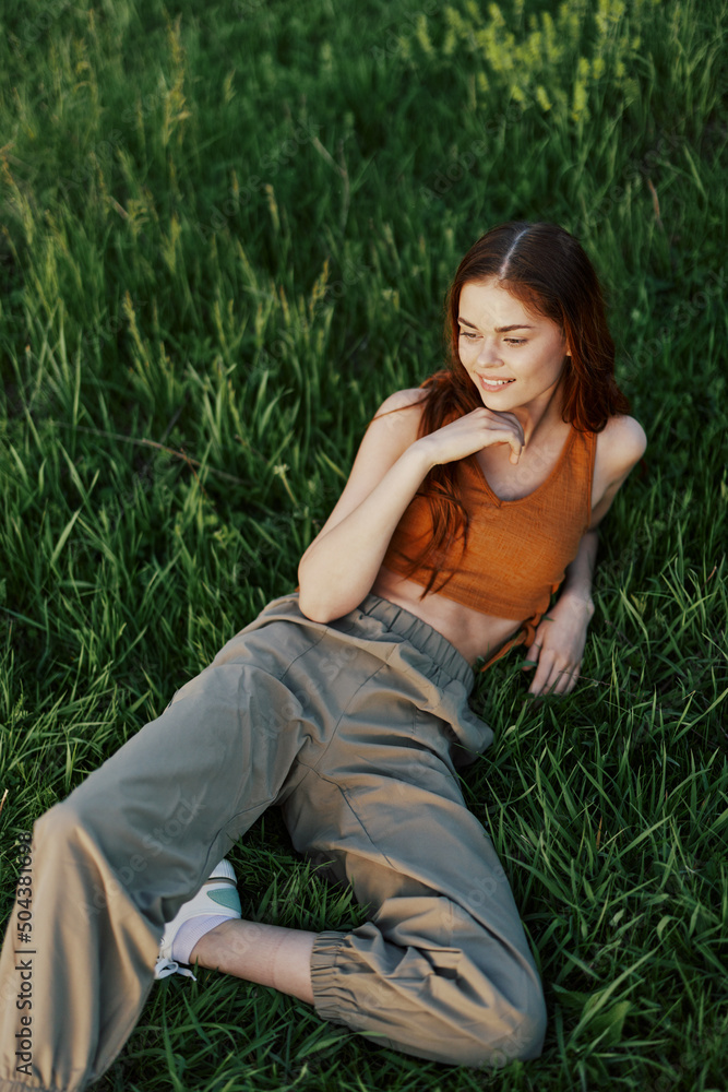 A woman lying on the green grass in green pants and an orange top, smiling in the sunset summer sun in nature A woman lying on the green grass in green pants and an orange top, smiling in the sunset summer sun in nature