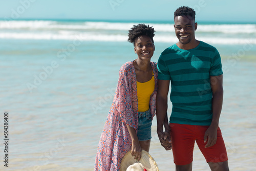 Portrait of happy african american young couple holding hands and standing against sea on sunny day
