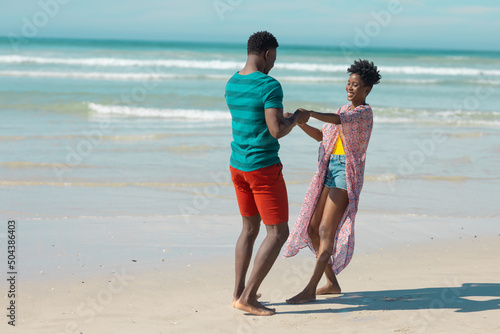 Cheerful african american young couple holding hands and dancing at beach against sky on sunny day