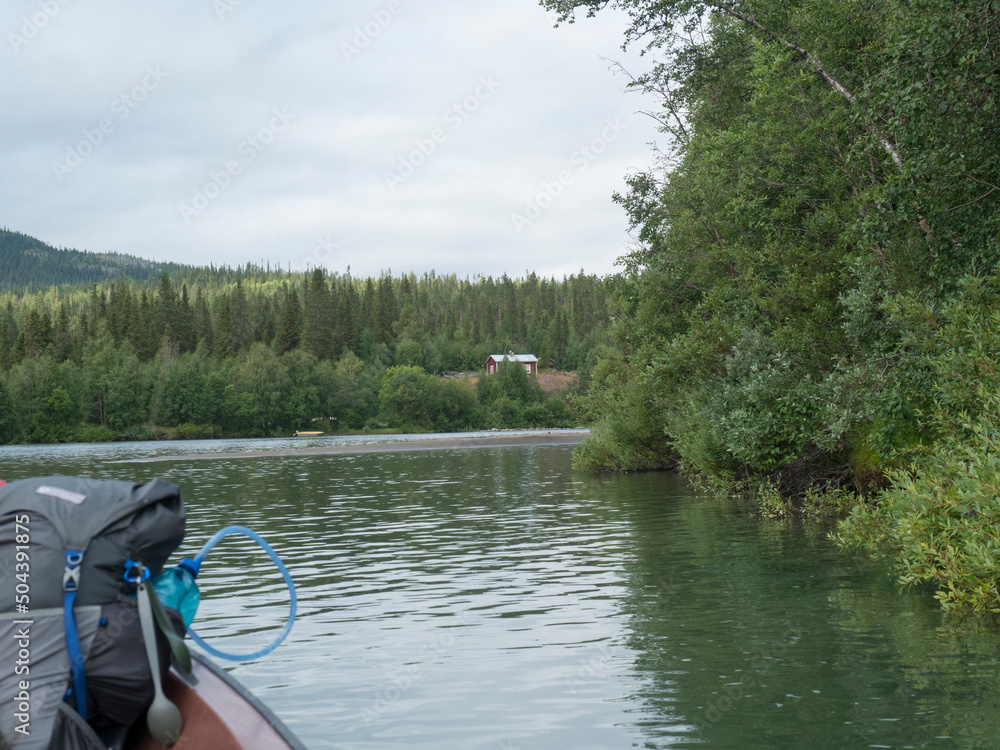 Fototapeta premium View of wide calm Tarra river from boat at Kvikkjokk, with forest and grass shore and green hills. Northern landscape in Swedish Lapland. End of Padjelantaleden hiking trail. Cloudy summer day