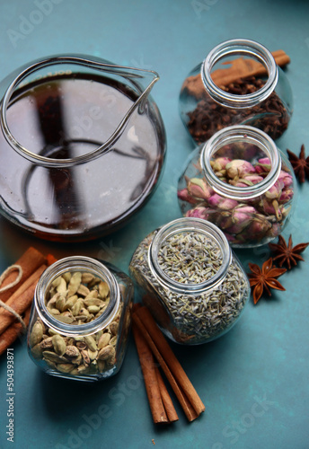 Teapot with black tea, glass jars with roses, lavender, clove, cardamom, anise stars and cinnamon sticks top view photo. Different types of tea spices on a table. Textured background with copy space. 