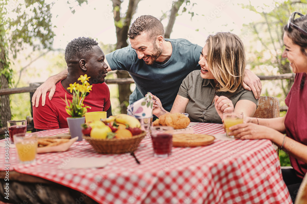 Multiethnic group of people gather on farm in the morning for breakfast picnic - friends sitting at an outdoor table drinking fruit juices and eating tarts and fruit