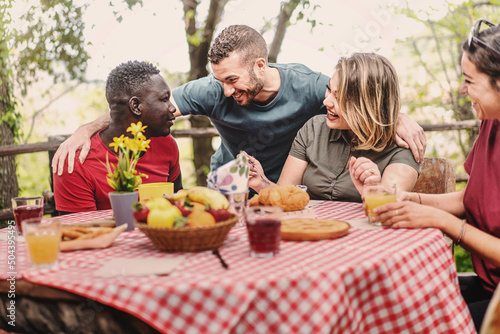 Multiethnic group of people gather on farm in the morning for breakfast picnic - friends sitting at an outdoor table drinking fruit juices and eating tarts and fruit