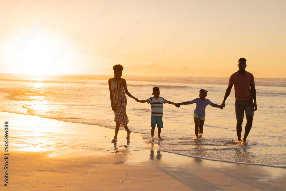 African american young parents holding son and daughter's hands while walking at shore against sky