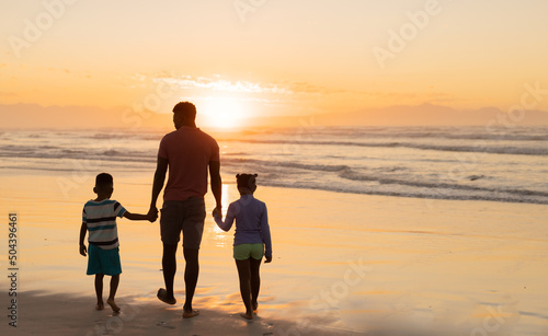 Rear view of african american young man holding son and daughter's hands while walking on beach